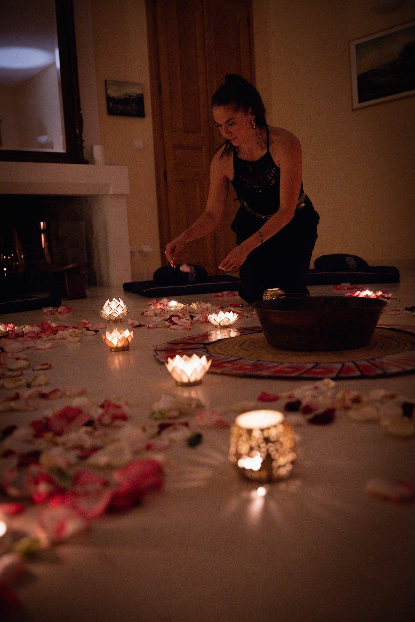 Susana à genoux dispose des pétales de rose autour de photophores lotus allumés, près d'un bol en cuivre, dans une pièce à la lumière tamisée
