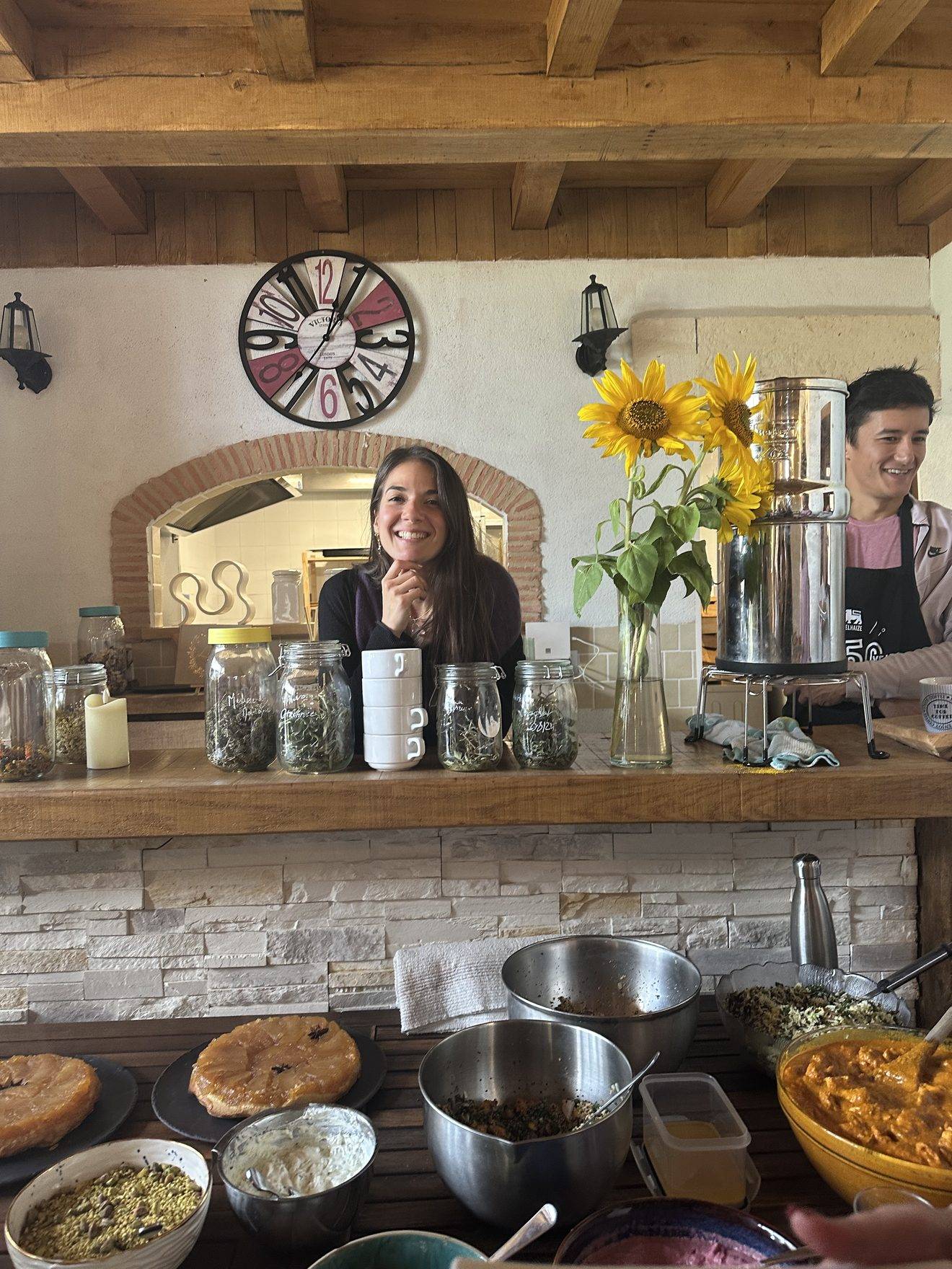 Susana d'Arcambel souriante derrière le comptoir de la cuisine, avec tournesols et plats préparés