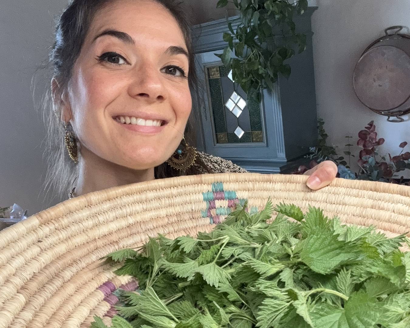 Susana souriante tenant un grand panier en osier tressé rempli d’orties fraîchement cueillies