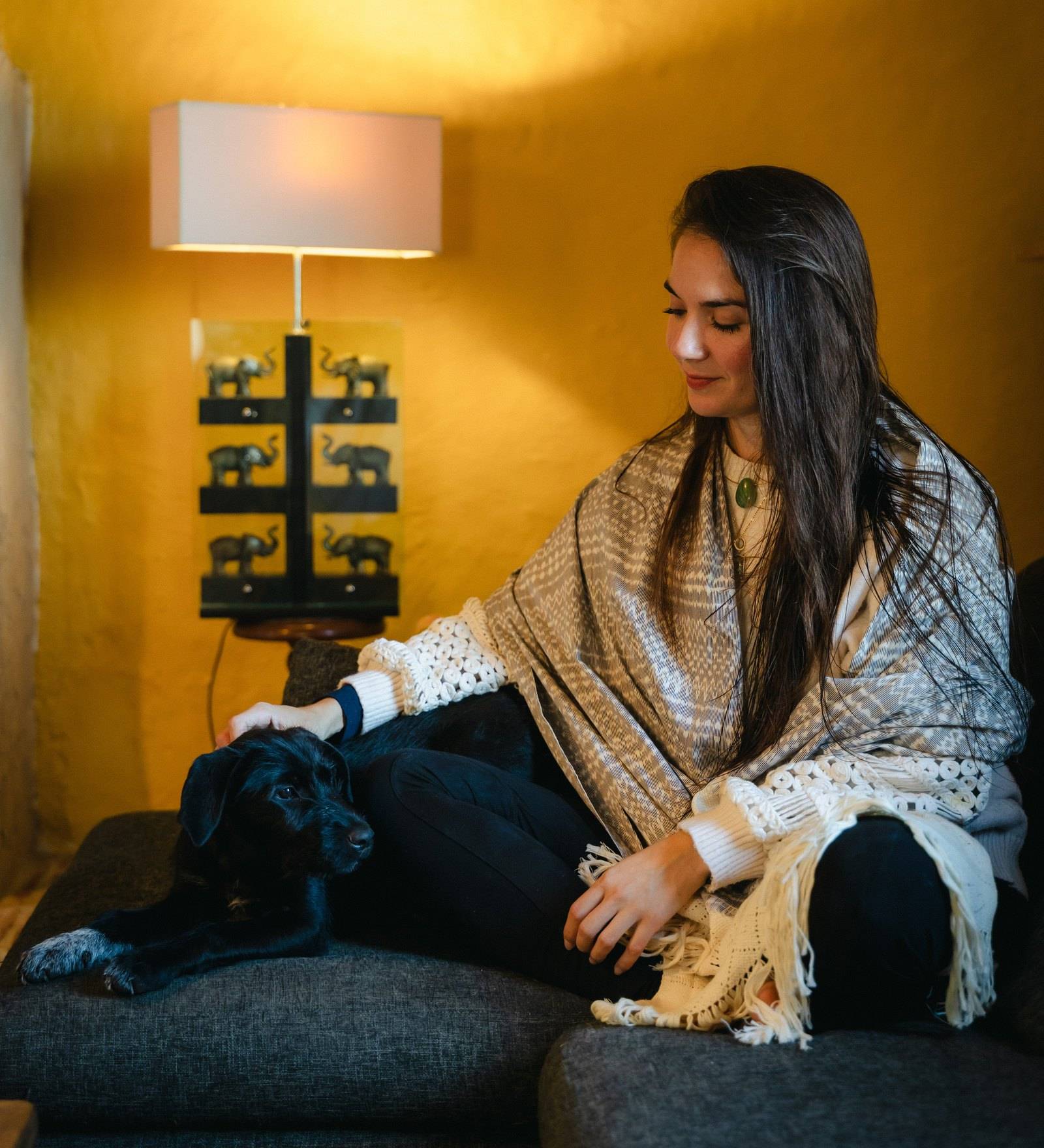 Susana d'Arcambel assise sur un canapé gris dans un salon aux murs ocre, un châle sur les épaules, sa chienne noire allongée à côté d'elle